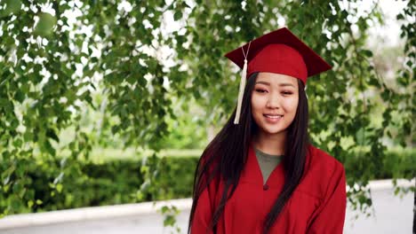 Portrait-of-attractive-Asian-girl-successful-graduating-student-in-gown-and-mortar-board-standing-on-campus,-smiling-and-looking-at-camera.-Youth-and-education-concept.