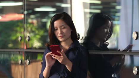 Young-Asian-Businesswoman-Touching-Mobile-Phone-Screen-Leaning-the-Office-Window-Glass,-4k