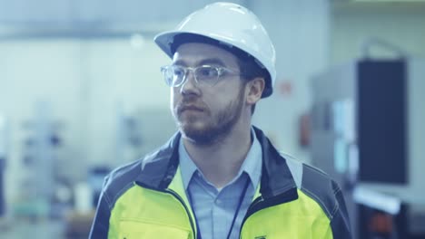 Industrial-Engineer-Wearing-Protective-Clothing-Puts-on-Hard-Hat-and-Walks-Through-Modern-Manufacturing-Facility-with-Automatic-Machinery-Working-in-Background.