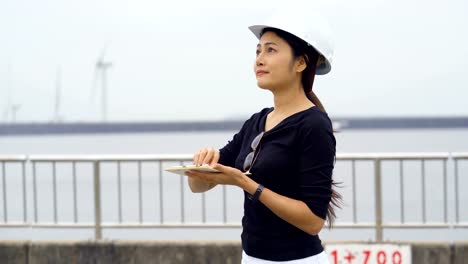 Asian-female-engineer-checks-job-The-background-is-a-large-reservoir-and-rotating-wind-turbines.