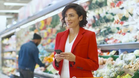 At-the-Supermarket:-Beautiful-Woman-Uses-Smartphone-while-Standing-in-the-Fresh-Produce-Section-of-the-Store.-Woman-Immersed-in-Internet-Surfing-on-Her-Mobile-Phone-In-the-Background-Colorful-Fruits-and-Organic-Vegetables.