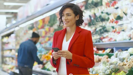 Im-Supermarkt:-schöne-Frau-nutzt-Smartphone-stehen-im-frisch-produzieren-Abschnitt-des-Speichers.-Frau,-die-mitten-im-Internetsurfen-auf-ihrem-Handy-im-Hintergrund-bunten-Obst-und-Bio-Gemüse.