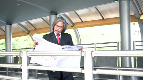 Senior-business-man-with-grey-hair-reading-newspaper-while-standing-and-waiting-at-train-station