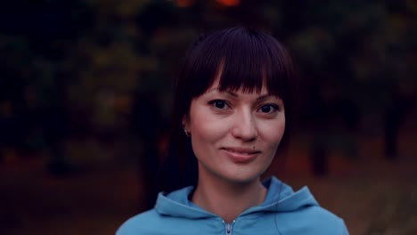 Close-up-portrait-of-beautiful-brunette-with-ponytail-wearing-bright-hoody-looking-at-camera-and-smiling-standing-in-the-park-in-the-evening-in-autumn.