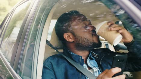 Negro-guapo-viajando-en-un-coche,-sentado-en-un-asiento-de-pasajero-utiliza-el-Smartphone,-toma-café.-Cámara-de-disparo-desde-fuera-del-vehículo.