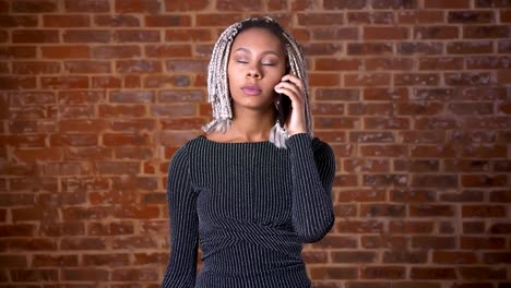 Young-African-girl-with-dreadlocks-talking-on-the-phone,-Brick-wall-in-the-background.