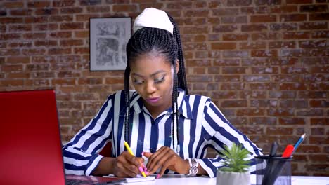 concentrated-african-woman-writting-notes-and-sitting-at-the-workplace-near-red-laptop-on-brick-background