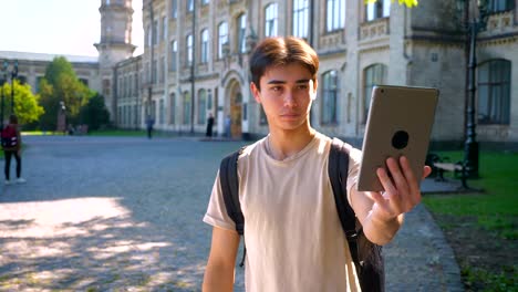 Glad-asian-male-is-taking-selfies-on-his-tablet-and-standing-straight,-near-school-building,-separated,-summertime