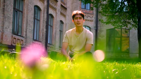 Calm-nice-asian-man-in-white-earphones,-concentrated-on-music-listening-and-having-rest-while-sitting-on-the-grass-in-park