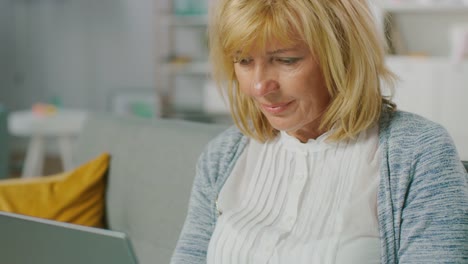 Portrait-of-a-Beautiful-Middle-Age-Woman-Relaxes-on-a-Sofa-in-Her-Living-Room,-Uses-Laptop.-Woman-Browses-Through-Internet-and-Doing-Work-from-Home-Office.