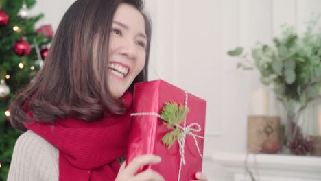 Cheerful-happy-young-Asian-woman-holding-christmas-gifts-smiling-to-camera-in-her-living-room-at-home-in-Christmas-Festival.-Lifestyle-woman-celebrate-Christmas-and-New-year-concept.