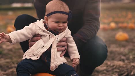 Close-up-of-a-baby-sitting-on-a-pumpkin-at-a-pumpkin-patch,-with-her-dad-holding-her-up