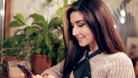 Young-brunette-woman-in-harness-at-cafe-using-mobile-phone,-gets-a-message-and-smiles.-Cup-of-hot-cacao-at-the-table.