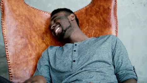 Portrait-of-African-man-sitting-in-chair,-using-Smartphone.-Man-reads-messages-and-laughs.-Guy-typing-massages,-smiling