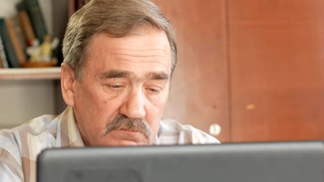 An-elderly-man-with-a-mustache-sits-behind-a-laptop-and-solves-problems.-He-looks-seriously-at-the-monitor