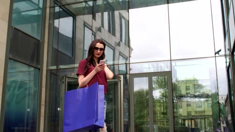 Chica-joven-con-teléfono-y-bolsas-cerca-de-centro-comercial.