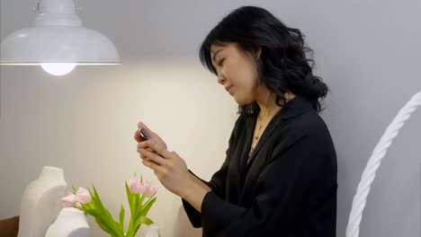 Asiatische-Frau-Smartphone-Bildschirm-betrachten-und-lächelnd-in-ein-Wohnzimmer