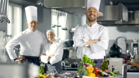 Famous-Chef-of-a-Big-Restaurant-Crosses-Arms-and-Smiles-in-a-Modern-Kitchen.-His-Staff-in-Smiling-in-the-Background.