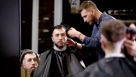 Stilvolle-Barber-ist-rasieren-Tempel-mit-Haarschneider-eines-bärtigen-Mannes-sitzt-auf-Sessel-im-Frisörsalon-und-Blick-auf-den-Spiegel.-Jungen-Coiffeur-schneidet-die-Haare-seines-Mandanten-in-den-schwarzen-Umhang