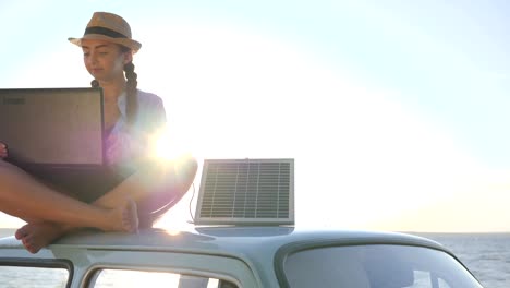 weekend-since-new-technology,-female-sitting-on-car-rooftop-with-notebook-and-battery-on-background-sky-in-backlit