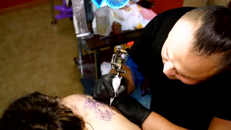 in-a-tattoo-salon-,-a-specialist-is-doing-a-tattoo-on-woman's-back,-a-floral-ornament.-a-man-works-in-special-gloves,-on-special-equipment.-drawing-is-done-with-black-paint