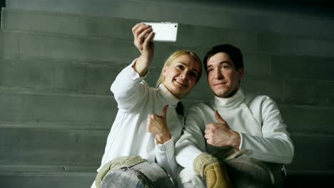 Two-young-smiling-fencers-man-and-woman-taking-self-portrait-on-smartphone-camera-after-fencing-training-indoors