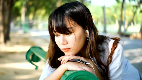 Beautiful-young-hipster-sad-asian-woman-emotionally-listening-music-in-headphones-with-smartphone,-sitting-on-the-park-bench-in-summer-sunset-sun-light.