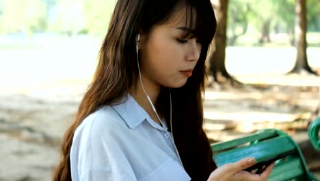 Linda-mujer-es-mensaje-de-texto-agradable-de-leer-en-el-teléfono-móvil-mientras-está-sentado-en-el-parque-en-primavera-cálida,-hermosa-mujer-es-escuchando-música-en-auriculares-y-buscando-información-sobre-el-teléfono-celular