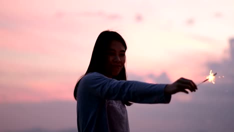 Young-asian-woman-stands-on-beach-with-sparkler-at-sunset-in-slow-motion.