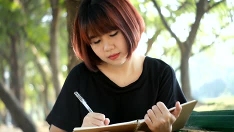 Hipster-feliz-joven-mujer-asiática-escribiendo-en-su-diario-en-el-parque.-Hipster-feliz-joven-mujer-asiática-trabajando-en-notebook-en-el-parque.-Estudiante-al-aire-libre.-Concepto-al-aire-libre-de-la-mujer-de-estilo-de-vida.