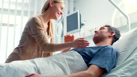 En-el-Hospital,-feliz-esposa-visita-a-su-marido-de-recuperación-que-es-mentira-en-la-cama.-Abrazar-con-amor-y-sonreír.