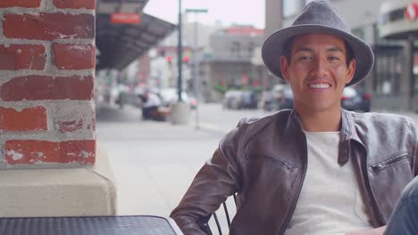 Young-man-sitting-outside-a-coffee-shop-smiles-and-puts-a-hat-on