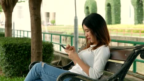 Young-asian-woman-using-smartphone-sitting-on-a-bench-in-a-public-park.