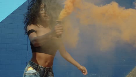 Beautiful-young-woman-holding-colorful-smoke-grenade-dancing-outside-against-blue-wall