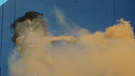 Beautiful-young-woman-holding-colorful-smoke-grenade-dancing-outside-against-blue-wall