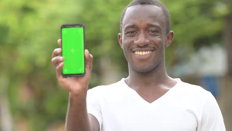 Joven-hombre-feliz-sonriendo-al-tiempo-que-muestra-el-teléfono-en-las-calles-al-aire-libre