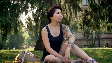 Trendy-young-asian-female-hipster-with-tattoo-sitting-on-grass-in-park-near-university,-thoughtful