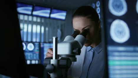 Científico-de-la-investigación-médica-femenina-mirando-a-través-de-los-tipos-de-microscopio-adquirió-datos-en-la-computadora.-Laboratorio.-En-el-laboratorio-con-múltiples-pantallas-MRI-/-CT-cerebral-escanear-imágenes.