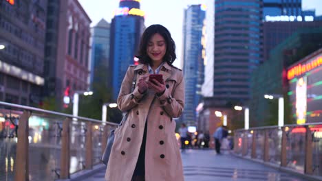 Slow-Motion-ziemlich-glücklich-junge-asiatische-Frau,-die-zu-Fuß-in-der-Stadtstraße-mit-Smartphone-am-Abend