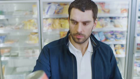 Portrait-Shot-of-the-Handsome-Man-Thinking-Very-Hard-and-Choosing-Tin-Can-from-the-Canned-Goods-Section,-Plaves-it-i-His-Shopping-Cart.-In-the-Background-Frozen-Goods-Section-of-the-Store.