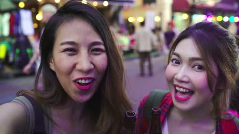 Traveler-backpacker-blogger-Asian-women-lesbian-lgbt-couple-travel-using-smartphone-for-selfie-in-Thailand.-Female-drinking-alcohol-or-beer-at-Khao-San-Road-the-most-famous-street-in-Bangkok.