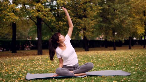 Beautiful-girl-is-concentrated-on-yoga-practice-sitting-on-mat-in-park-in-lotus-position-and-bending-sideways-raising-arm.-Healthy-lifestyle-and-recreation-concept.