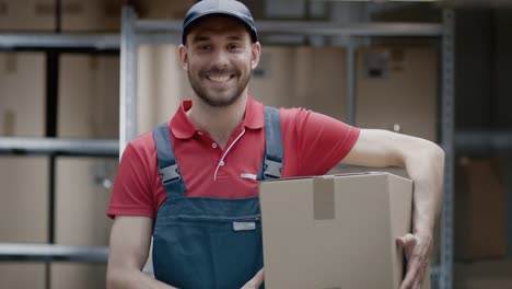 Portrait-of-Handsome-Warehouse-Worker-in-Uniform-Holds-Cardboard-Box-Package-and-Smiles.