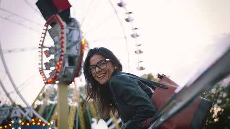 Mujer-joven-con-estilo-en-gafas-sonriendo-alegremente-delante-de-una-noria-en-el-parque-de-atracciones