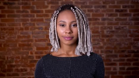 Young-African-girl-with-dreadlocks-smiling-and-looking-at-camera,-Brick-wall-in-the-background.