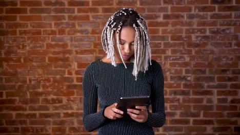 Young-African-girl-with-dreadlocks-using-a-tablet-computer,-typing.-Brick-wall-in-the-background.