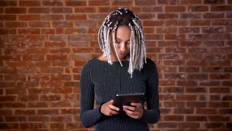 Niña-africana-con-rastas-usando-un-tablet-PC,-escribiendo.-Pared-de-ladrillos-en-el-fondo.