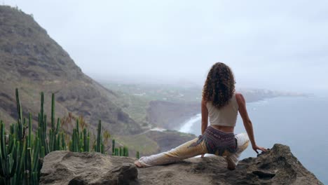 A-woman-sitting-on-the-edge-of-a-cliff-in-a-pose-war-overlooking-the-ocean-raise-her-hands-up-and-inhale-the-sea-air-while-doing-yoga