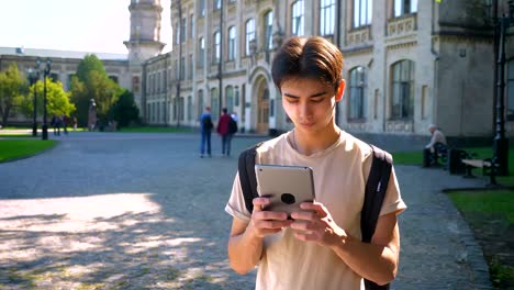 Konzentrierte-nette-asiatische-junge-nutzt-seinen-Tablet-stehend-in-erstaunliche-Altstadt,-Flecken-des-Sonnenlichts-in-Sommerlaune