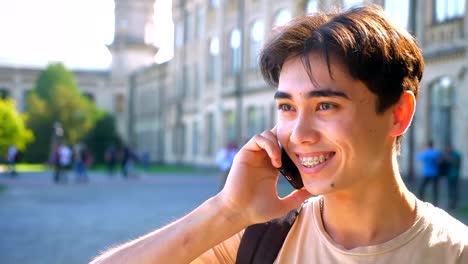 Nice-asian-boy-is-on-the-phone,-smiling-and-talking,-bright-sun,-urban-view-as-background